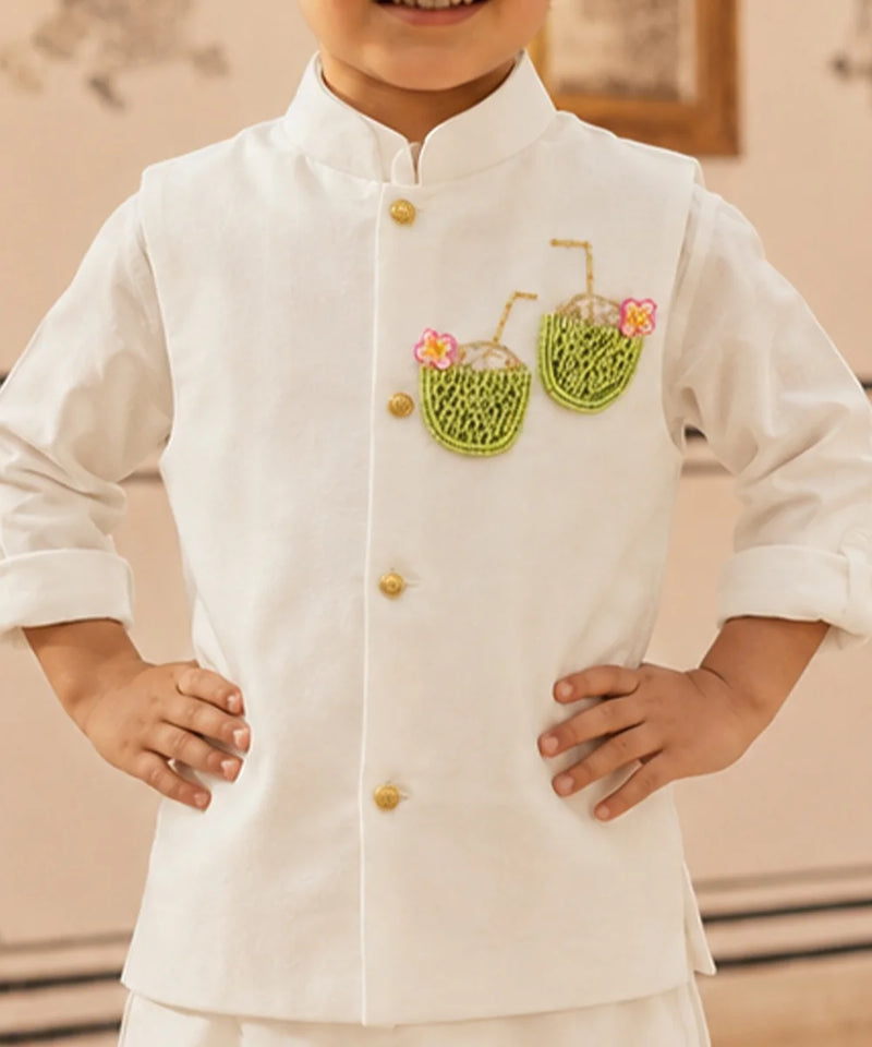 A child wearing a white traditional-style jacket with gold buttons and embroidered green coconut motifs with pink flowers on the chest, standing with hands on hips.