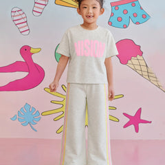 A young girl stands smiling in a matching light gray two-piece lounge set with pink and yellow accents. The set includes a short-sleeve t-shirt with 'VISION' printed in pink across the chest and matching wide-leg sweatpants with yellow side stripes.