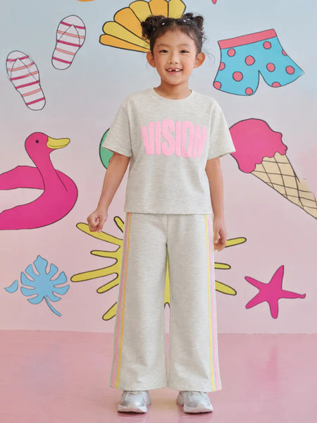 A young girl stands smiling in a matching light gray two-piece lounge set with pink and yellow accents. The set includes a short-sleeve t-shirt with 'VISION' printed in pink across the chest and matching wide-leg sweatpants with yellow side stripes.