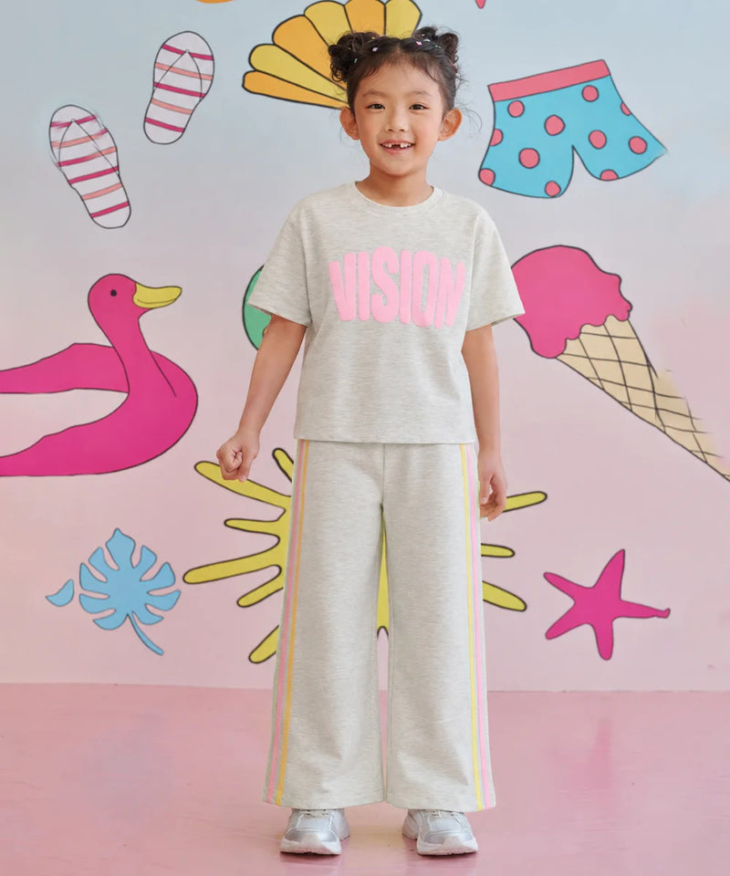 A young girl stands smiling in a matching light gray two-piece lounge set with pink and yellow accents. The set includes a short-sleeve t-shirt with 'VISION' printed in pink across the chest and matching wide-leg sweatpants with yellow side stripes.