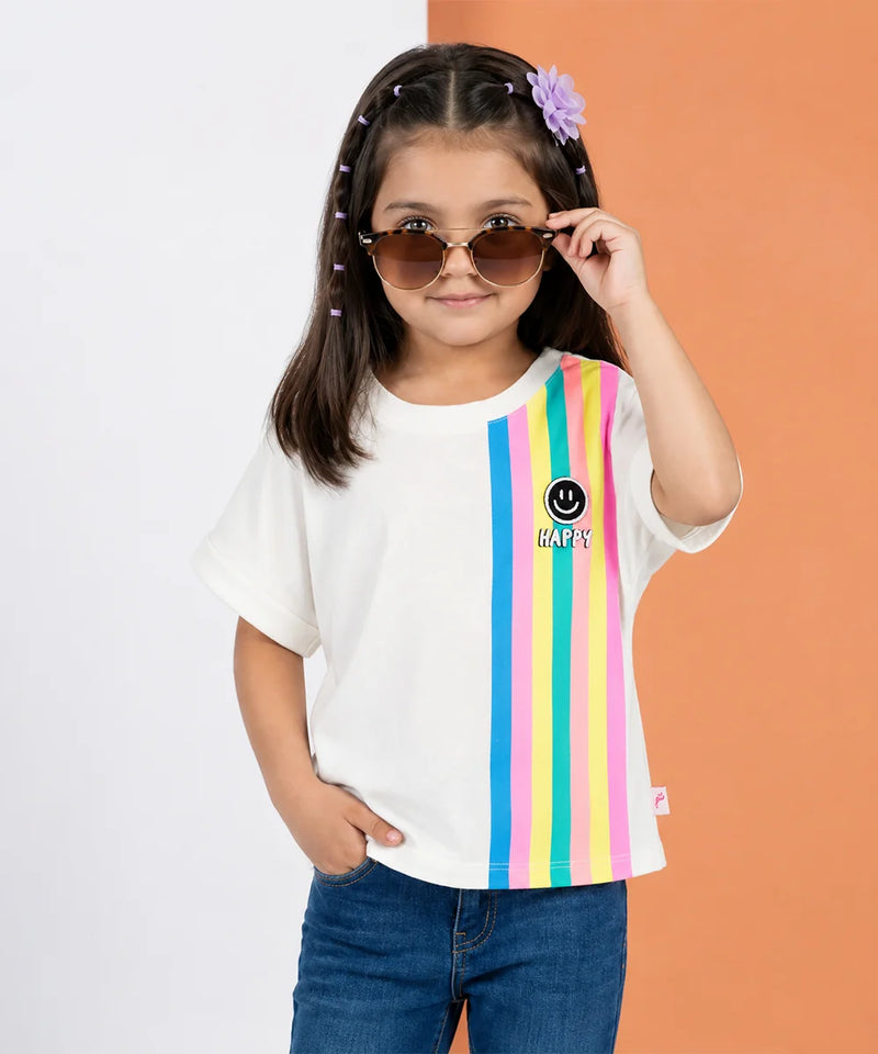 A young girl with sunglasses and a purple flower hair clip wears a white t-shirt with vertical rainbow stripes on the right side and a smiley face logo with 'KAPY' text. She is paired with blue jeans, standing against a split white and orange background.