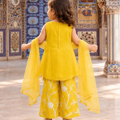 A young girl viewed from behind wearing a bright yellow traditional outfit with wide-leg embroidered pants and a matching sheer dupatta, standing in an ornate decorated interior.