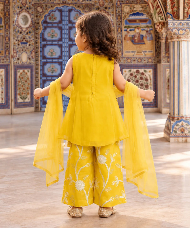 A young girl viewed from behind wearing a bright yellow traditional outfit with wide-leg embroidered pants and a matching sheer dupatta, standing in an ornate decorated interior.
