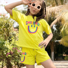 A young girl posing outdoors in a bright yellow two-piece lounge set featuring a smiley face graphic and the word 'SUMEBIRD' printed on the shirt and shorts.