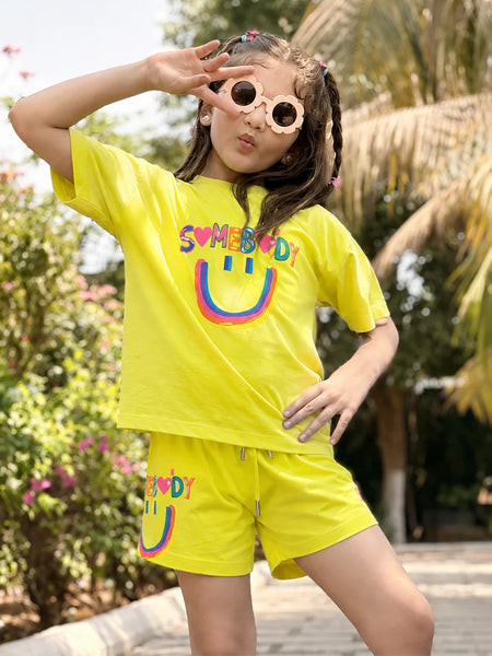 A young girl posing outdoors in a bright yellow two-piece lounge set featuring a smiley face graphic and the word 'SUMEBIRD' printed on the shirt and shorts.