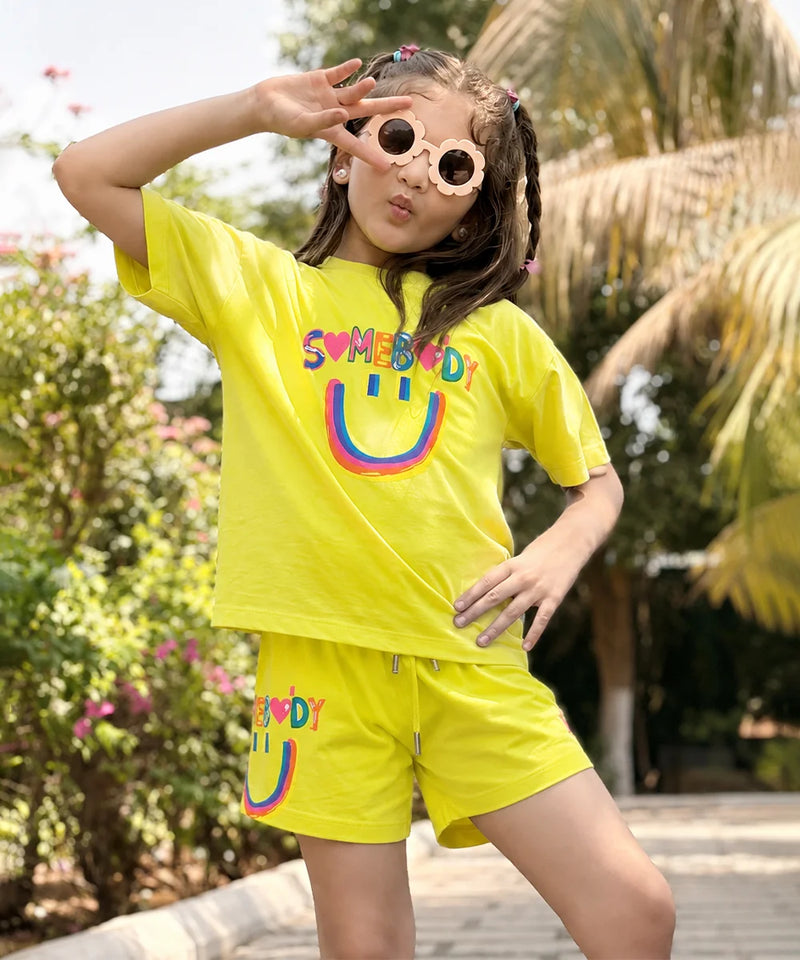 A young girl posing outdoors in a bright yellow two-piece lounge set featuring a smiley face graphic and the word 'SUMEBIRD' printed on the shirt and shorts.